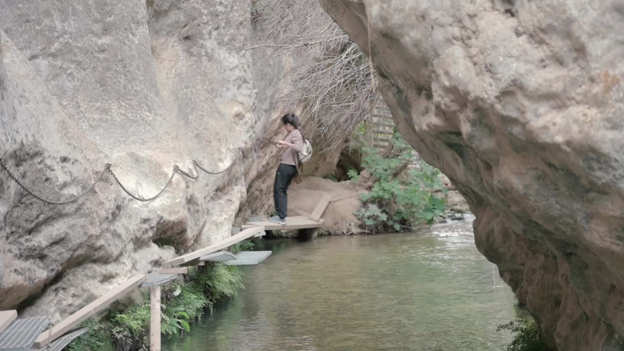 Hiker going across a rocky riverbed, holding onto a chain for support while walking on narrow, precarious wooden walkways