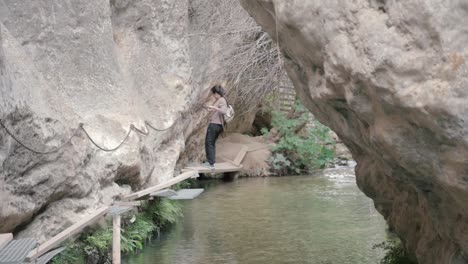 hiker going across a rocky riverbed, holding onto a chain for support while walking on narrow, precarious wooden walkways