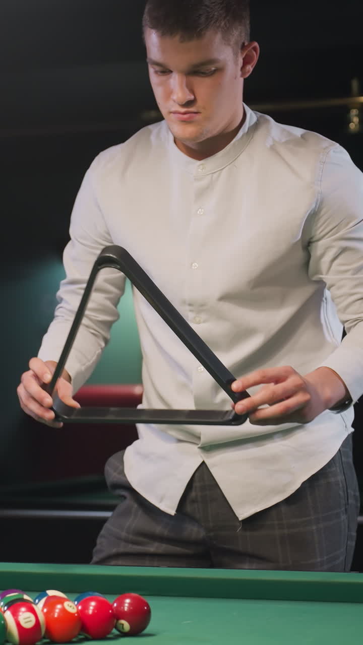 un aprendiz de billar con camisa blanca y pantalones grises arregla bolas de billar de colores en formación de triángulo en una mesa de billar verde vibrante bajo una cálida iluminación ambiental con una expresión facial seria