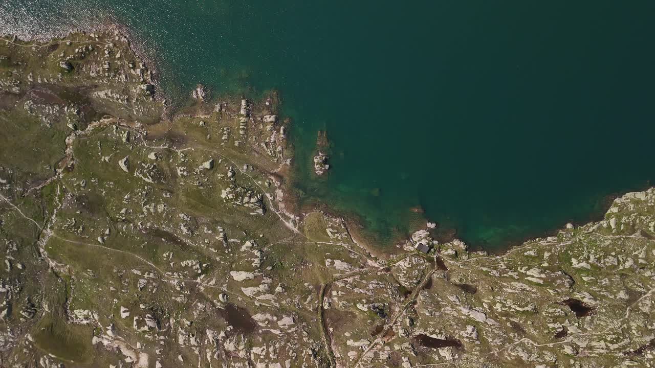 Top-down aerial view of a rugged lakeside bank, unveiling the clear waters of Lake Totensee in Obergoms, Switzerland, showcasing the natural beauty and dramatic contrast of the alpine landscape.