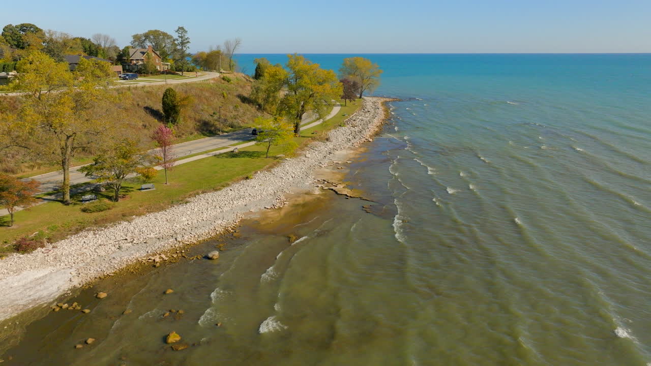 Drone aerial flying above Lake Michigan shoreline in Sheboygan, Wisconsin with cars along the road, colorful autumn trees, and neighborhoods on a bright, pretty day