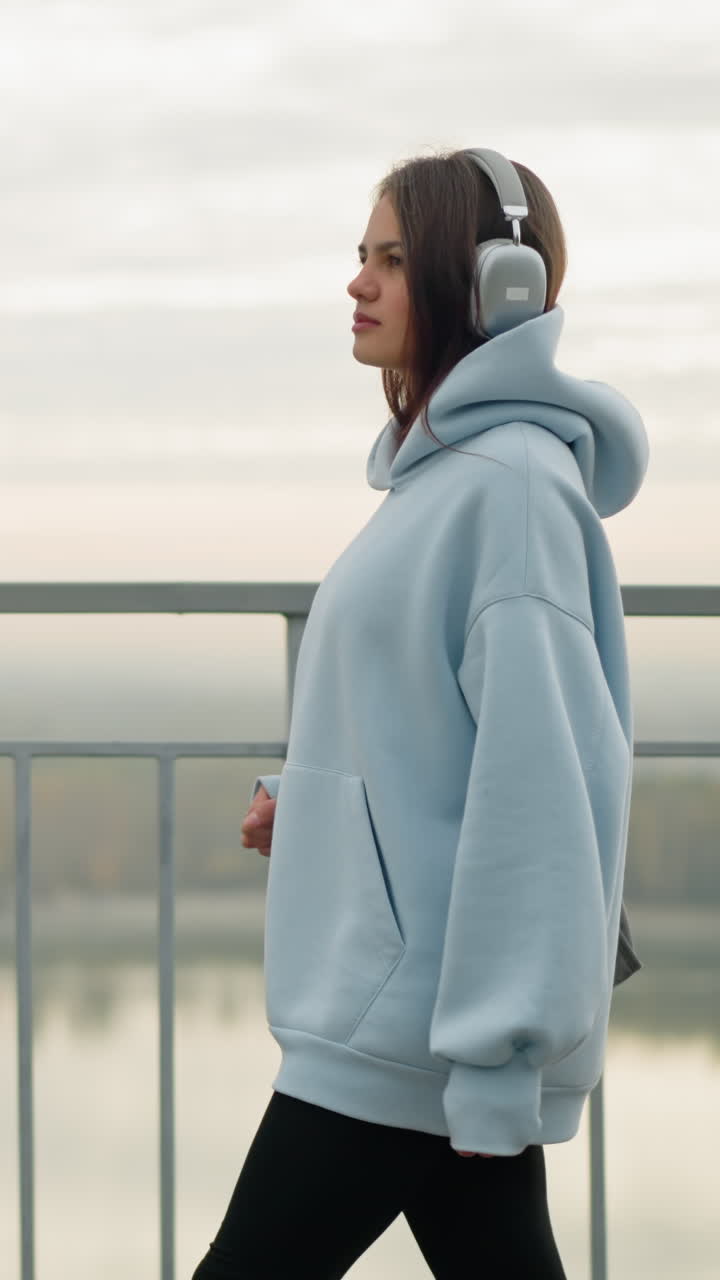 Vista laterale di una donna adulta che cammina lentamente mentre ascolta musica con le cuffie, godendosi una tranquilla passeggiata all'aperto in un contesto urbano, con un'atmosfera calma e dintorni sereni vicino al fiume e alla ringhiera.