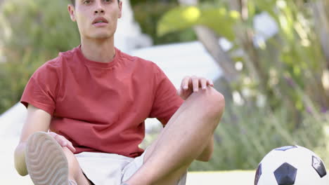 focused biracial man sitting with football, touching toes in sunny garden, copy space, slow motion