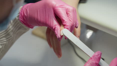 close-up view of nail technician wearing pink gloves carefully filing customer fingernail using nail file. technician holds client's hand gently while shaping nail in clean, well-lit salon environment