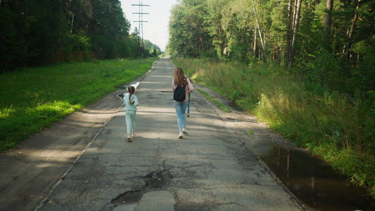 Energetic little boy runs ahead of mum and sister on cracked countryside road surrounded by lush green trees and puddles, capturing playful moment of freedom and joy in peaceful forest path