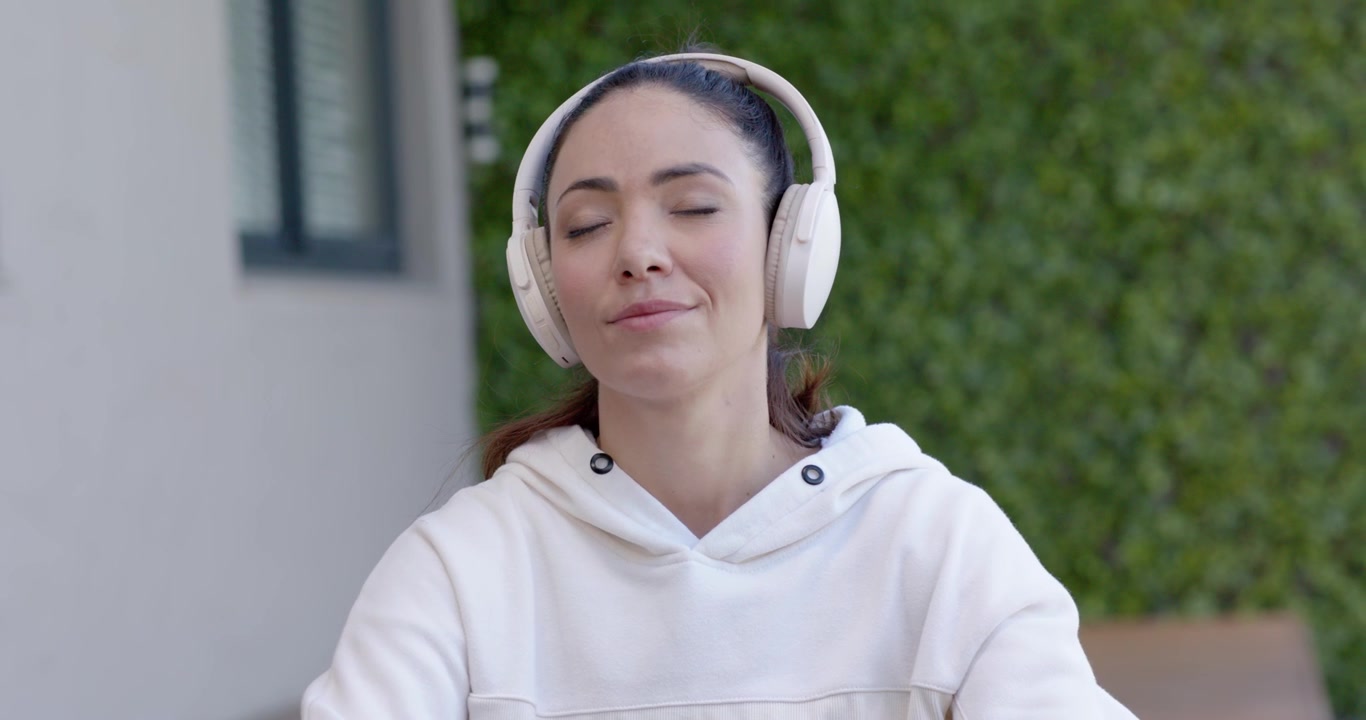 Donna con felpa con cappuccio che controlla il telefono rosso, chiudendo gli occhi, ascoltando con le cuffie su una panchina vicino a una parete vegetale.