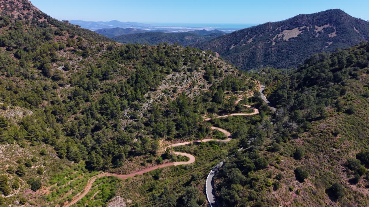 Strada di montagna tortuosa che si snoda tra i pendii boscosi della Sierra de Espadan, Spagna, con una costa lontana e il mare blu visibile all'orizzonte.