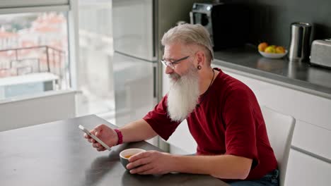 an elderly man with gray hair, a lush beard, glasses and a red t-shirt looks at his social media feed and drinks tea during his breakfast in the morning in a modern apartment overlooking the sea