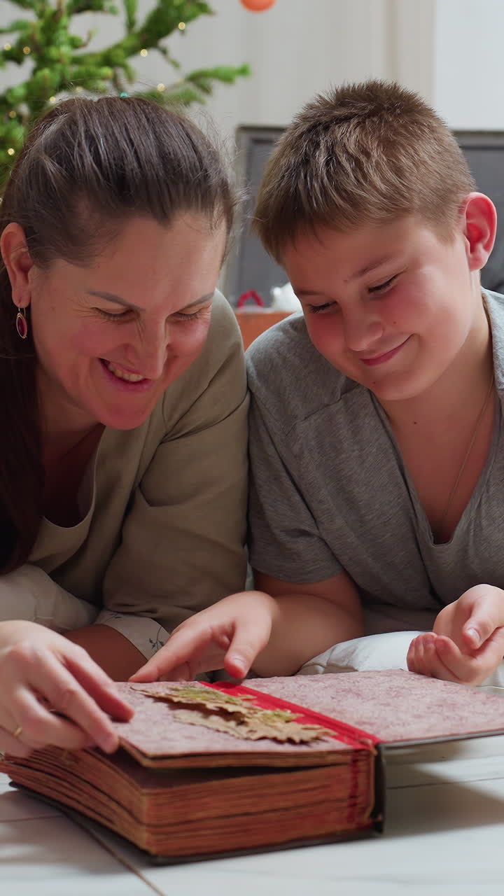 Madre e hijo blancos leyendo un álbum junto al árbol de Navidad decorado, hojeando páginas de fotos familiares antiguas, iluminación cálida, recuerdo acogedor de las fiestas, la madre narradora guía al niño curioso.