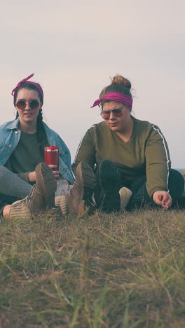 happy young women with hard drinks point to clear sky sitting on grass at tent in autumn evening low angle shot