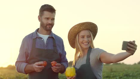 Alegres-Jóvenes-Y-Atractivos-Granjeros-Sonriendo-A-La-Cámara-Del-Smartphone-Y-Tomando-Fotos-Selfie-En-El-Campo-Con-Verduras-En-Las-Manos,-Y-Divirtiéndose-Con-Ellos