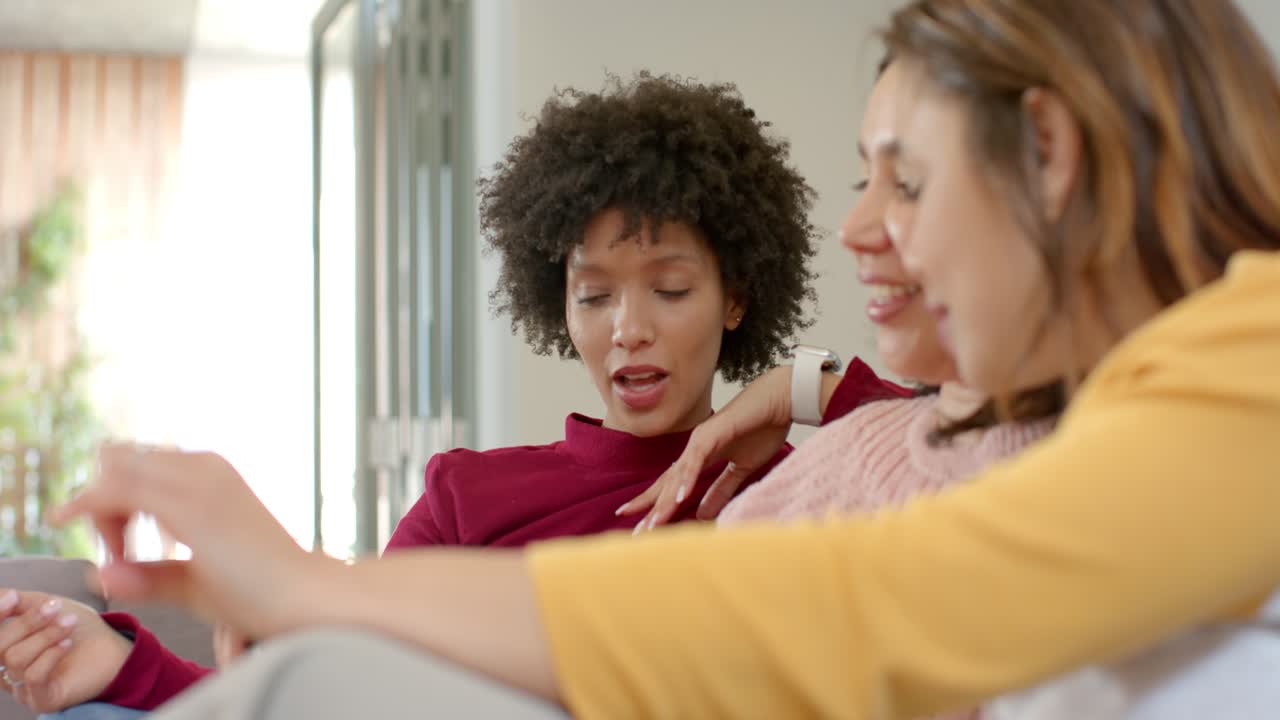 Diverse female friends on living room sofa gesturing, passing remote to change channel