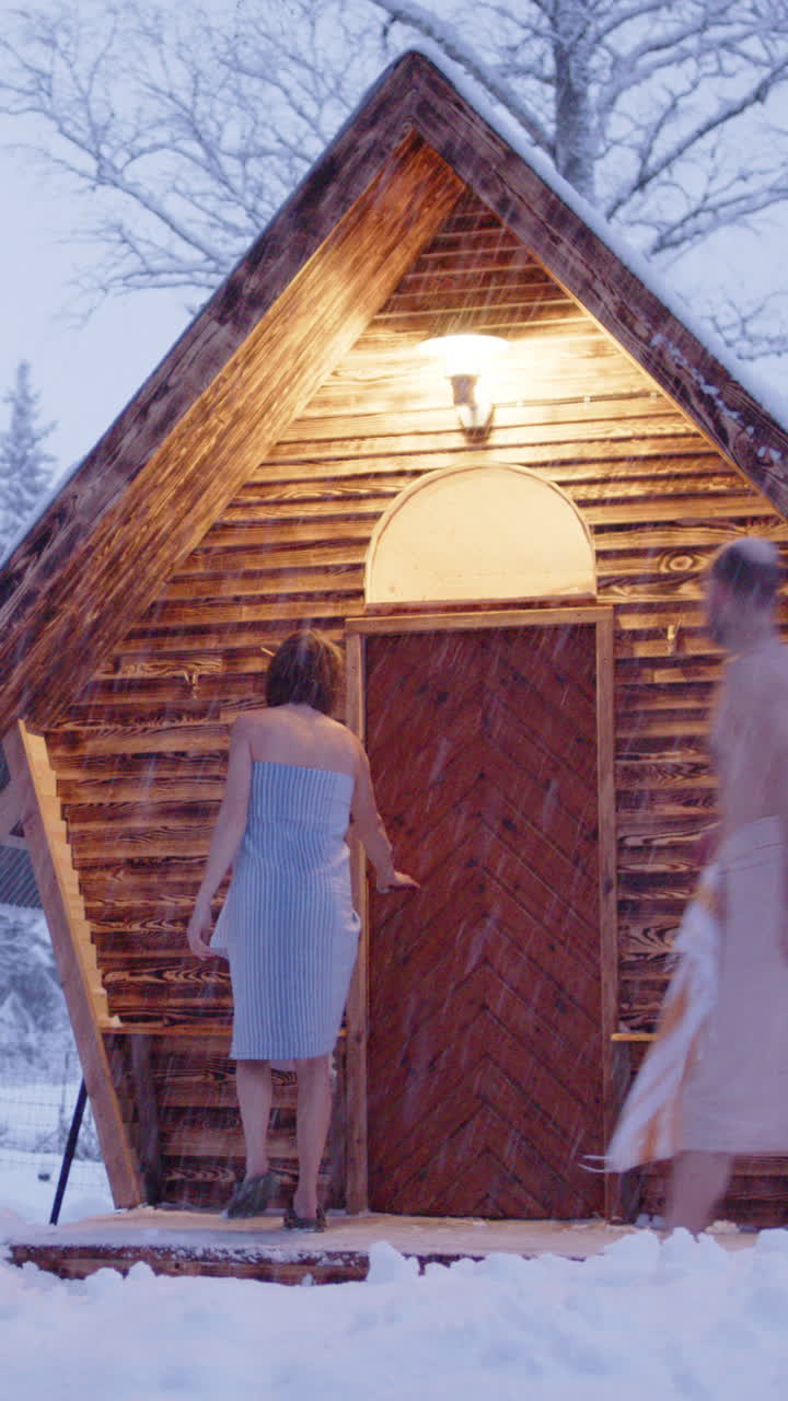 Scandinavian couple in towels enter wooden sauna in snowy woods. Vertical