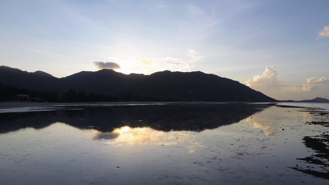 Drone footage capturing sunrise over Koh Phangan Thailand with the dark silhouette of a mountain and the reflection of sunlight shimmering on the calm tropical sea