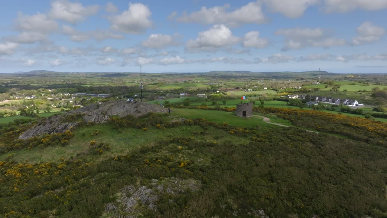 Vinegar Hill, Enniscorthy, County Wexford, Ireland, April 2025. Drone establishing orbit from rocky cliff with people sitting and admiring view to ascend above walking path to stone tower monument.