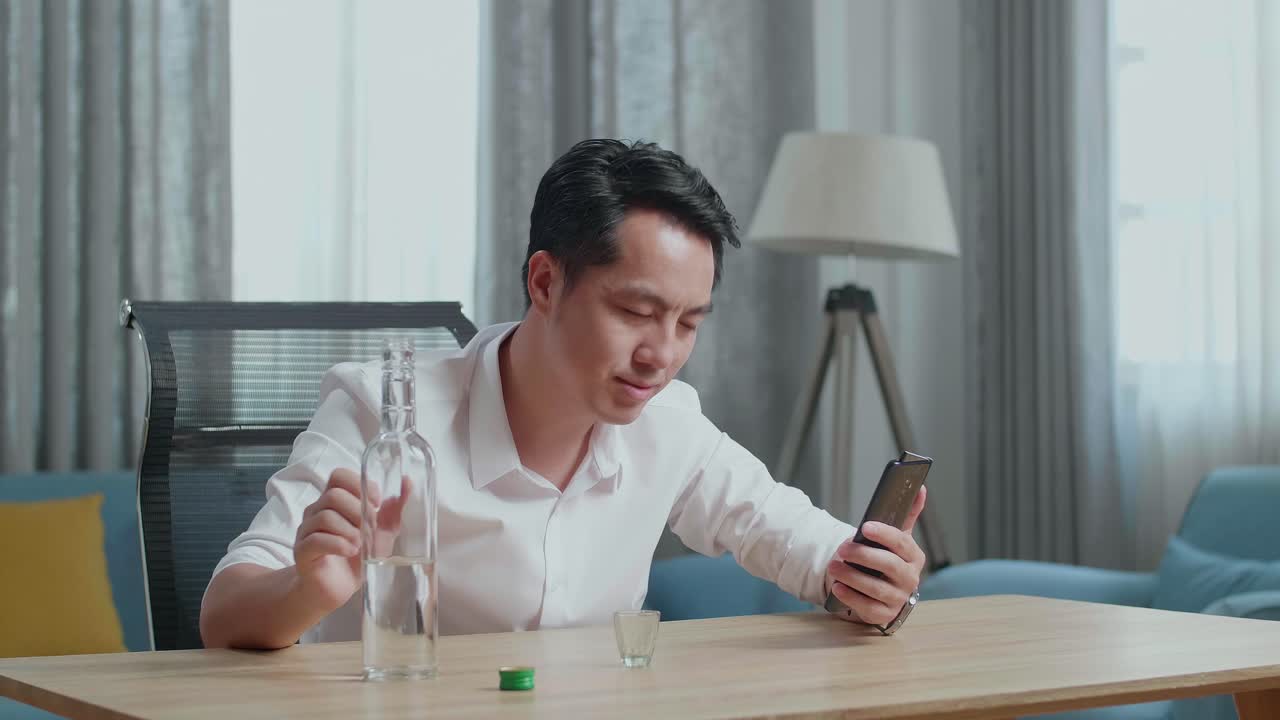 아시아인 남자가 집에서 스마트폰으로 비디오 통화를 하는 동안 환호하고 술을 마시기 전에  글래스에 보드카를 고 있습니다.