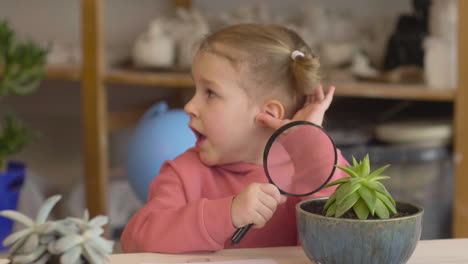 Little-Girl-Observing-A-Plant-With-A-Magnifying-Glass-Sitting-At-Table-In-A-Craft-Workshop