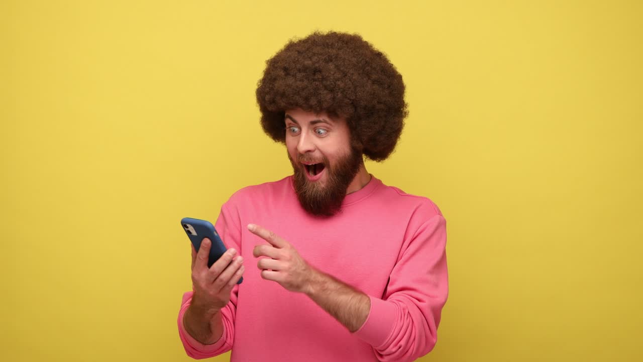 Bearded hipster man pointing at mobile phone with blank screen, surprised facial expression.