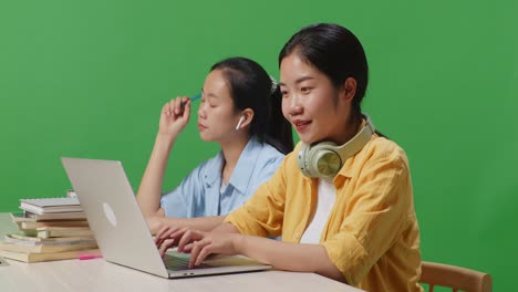 close up of woman student typing on a laptop while sitting with her friend studying on a table in the green screen background classroom