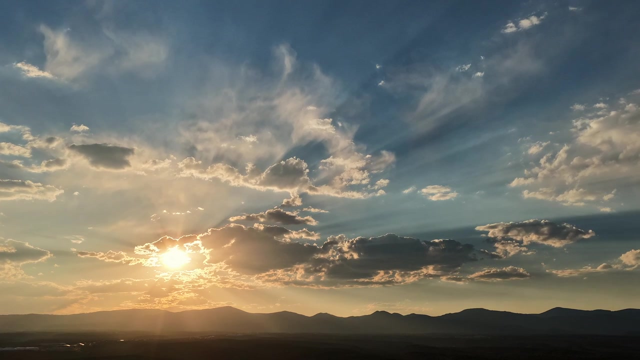 Un timelapse con drone 4K di decine di raggi di luce che si muovono attraverso un cielo brillante dorato e blu, enfatizzando modelli meteorologici drammatici su una catena montuosa del sud-ovest scura e in silhouette