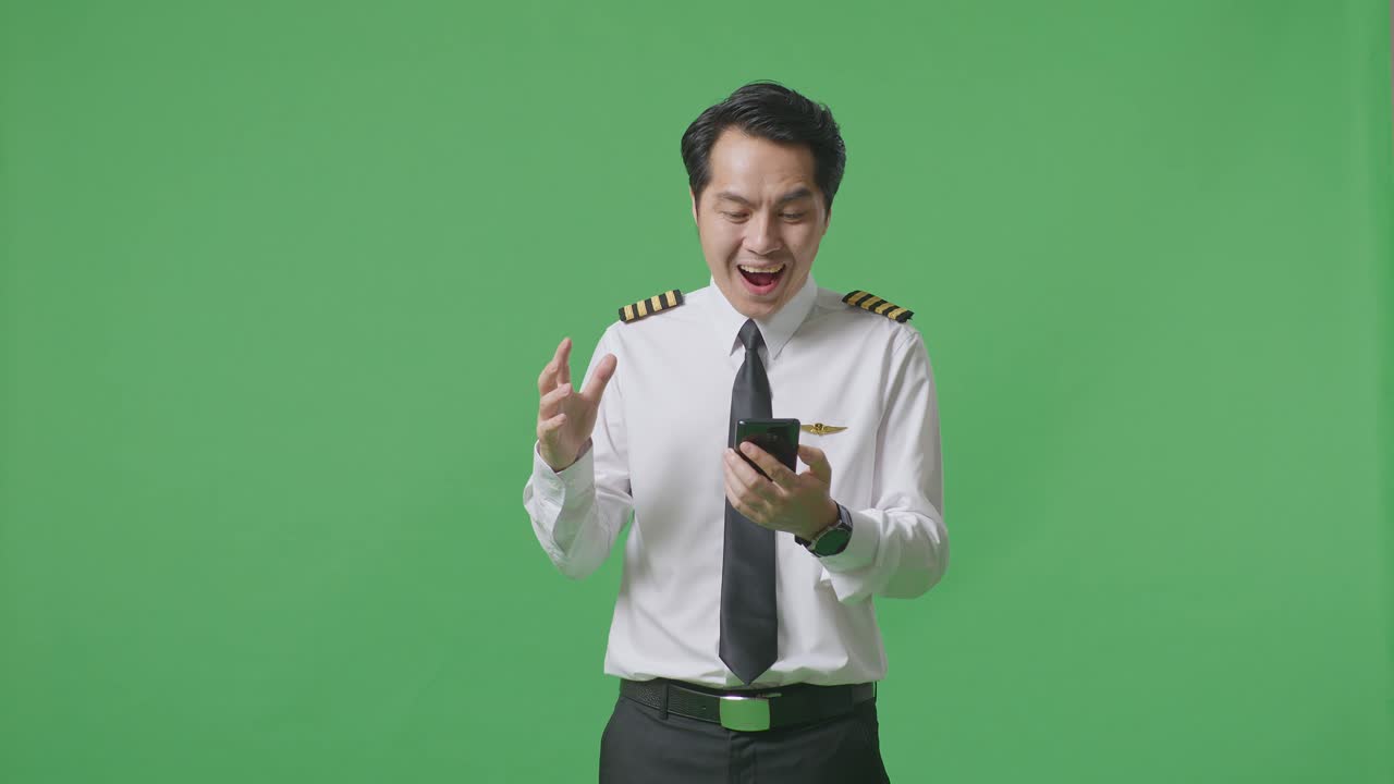 아시아인 조종사가 스마트폰을 보고 미소 짓고 초록색 스크린 배경 스튜디오에 서서 와우라고 말합니다.