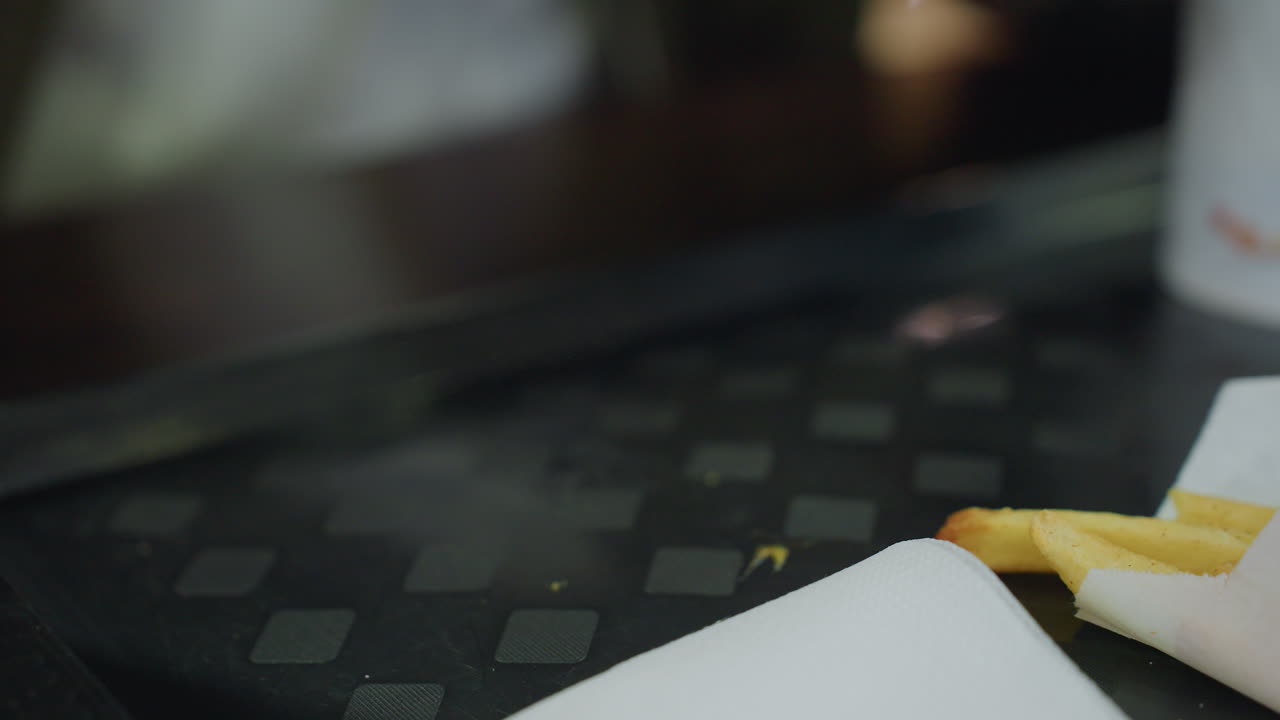 Primo piano di una mano scura che si allunga per prendere un succulento hamburger con pane ai semi di sesamo, formaggio fuso, ketchup e patatine fritte sul tavolo, in un ambiente informale che cattura lo stile di vita del fast food