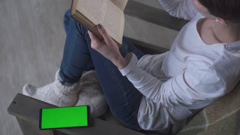 niña con cabello corto en vaqueros se sienta en casa en un sillón y lee un libro en invierno. cerca de teléfono móvil con pantalla verde. mujer con teléfono. teléfono celular femenino. llave de croma teléfono celular pantalla verde.