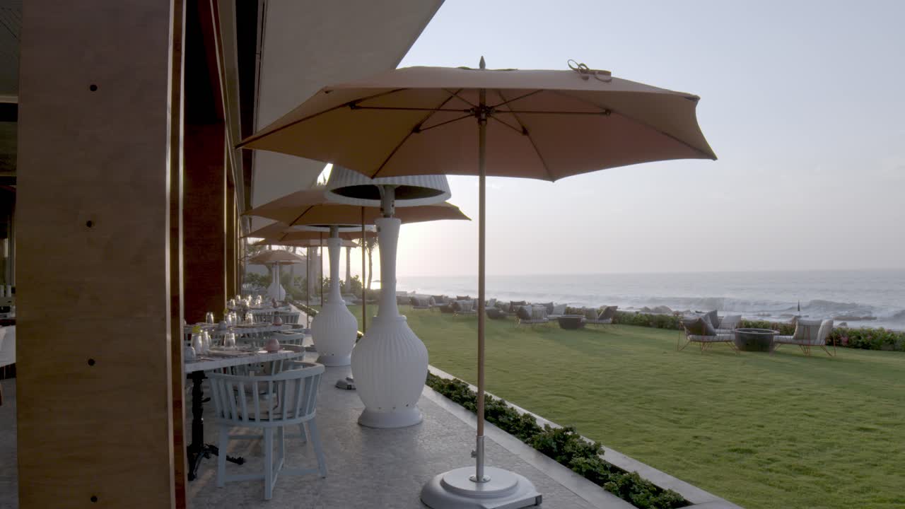 A wide shot captures a serene oceanfront resort patio with shaded tables, heaters, and lush green lawns, showcasing a relaxing vacation setting by the waves.