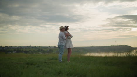 a tender moment between two sweethearts, as a man in a hat, white shirt, and jeans holds a woman in a white dress on a grassy hill at sunset. the scene exudes warmth, love, and connection