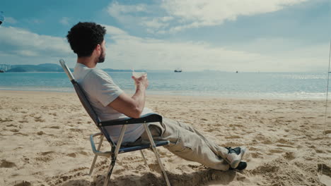 Slow motion shot of a man sipping caipirinha on a Brazilian beach, sitting in a folding chair with boats and mountains in the distance