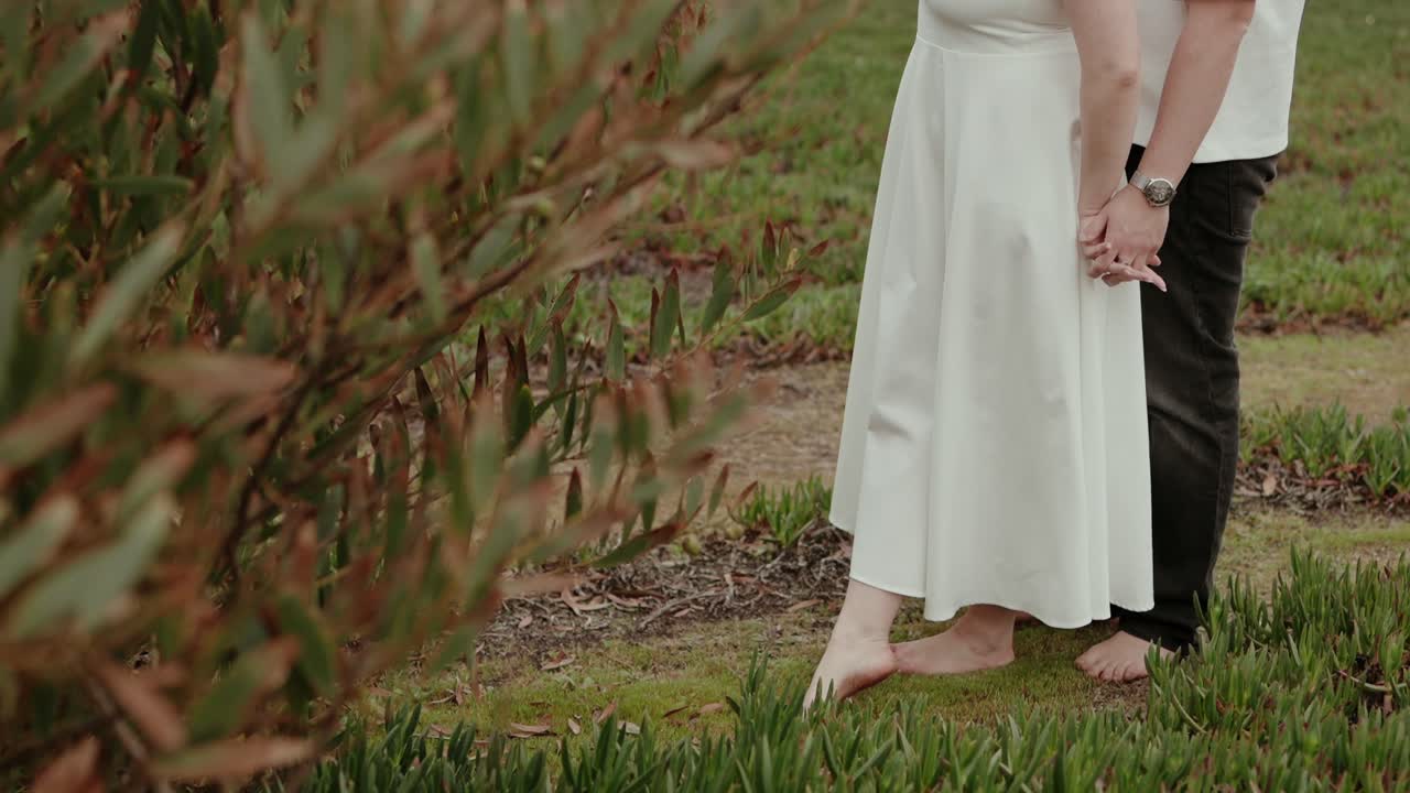 Lower body view of couple holding hands while standing barefoot on grass near plants in natural outdoor setting