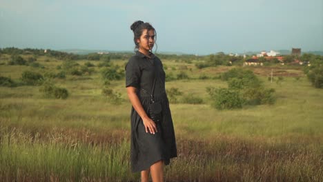 Pan-slow-motion-shot-of-an-Indian-model-in-a-black-dress-stading-in-the-middle-of-a-grassfield-during-sunset