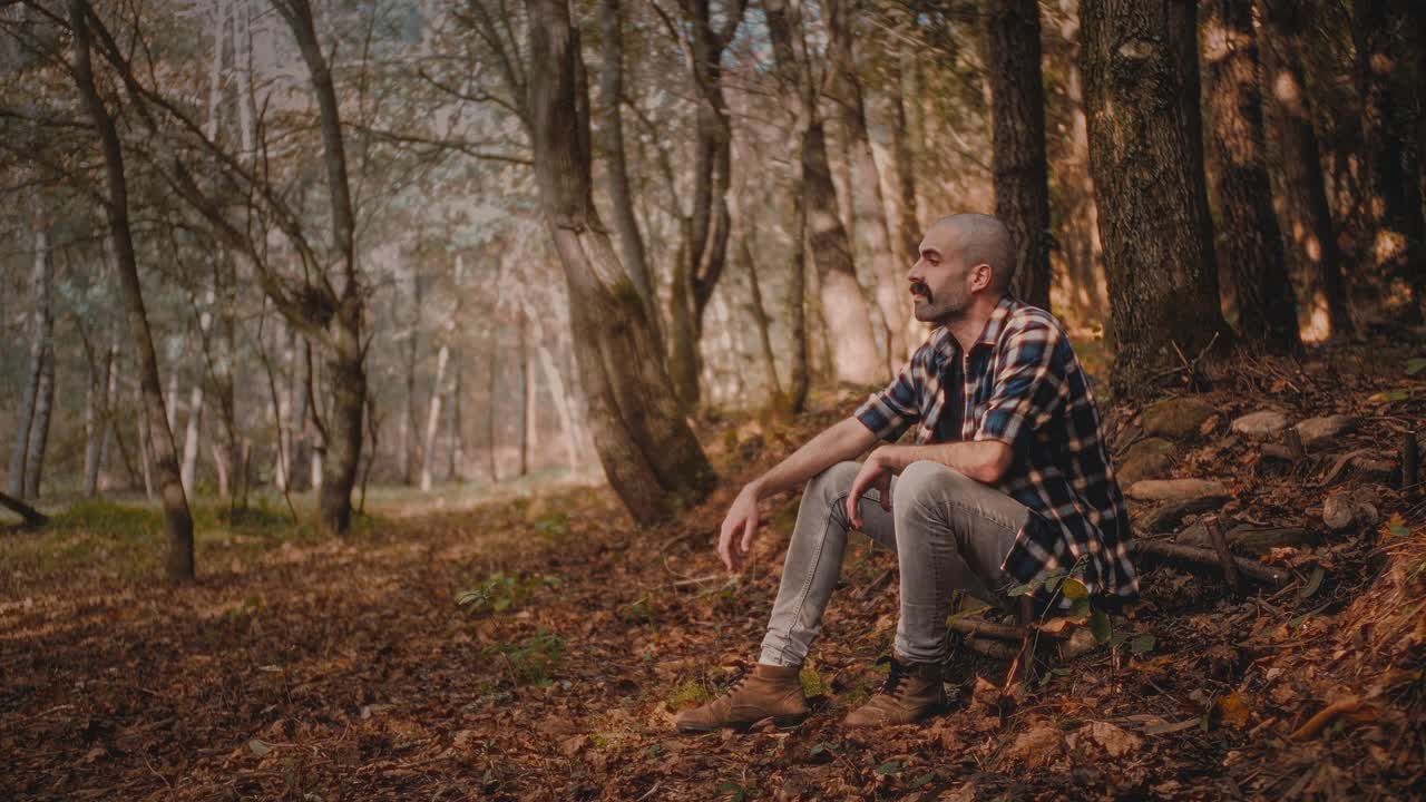 Young hipster in blue shirt sits on wooden stair in forest. He marvels at nature’s beauty. Warm and relaxing clip