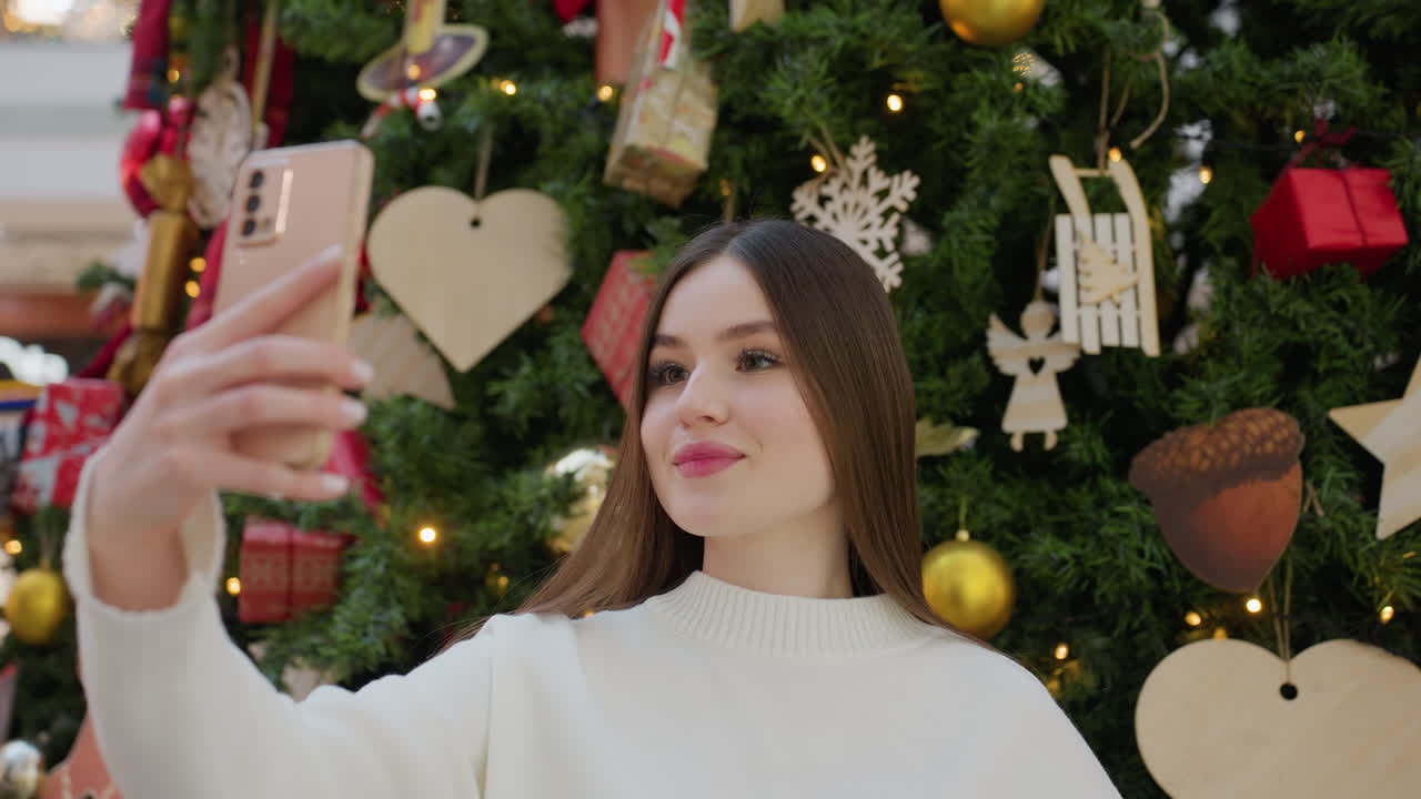 Young lady in white top posing for a selfie with decorative Christmas tree behind her, she strikes a playful pose with hand on head and bokeh light effect in the background