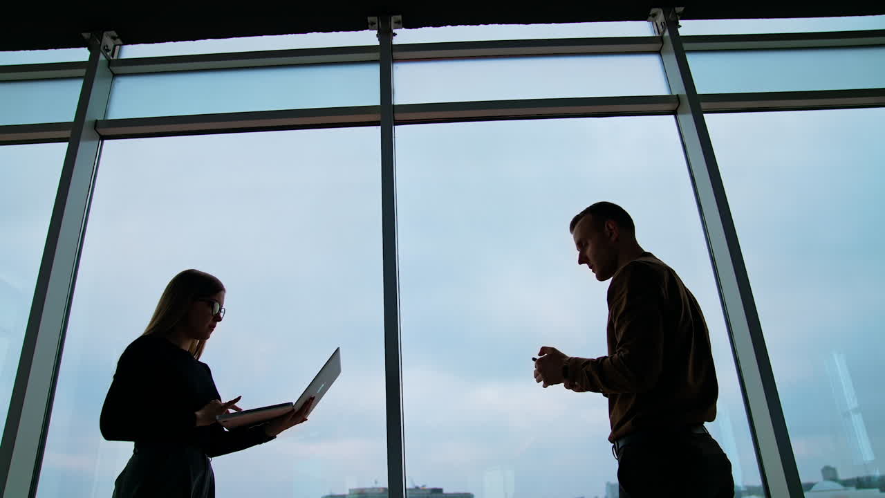 Young people work in office near large windows. Silhouette of business people on windows background in the evening. Woman uses a laptop and man a mobile phone in business center.