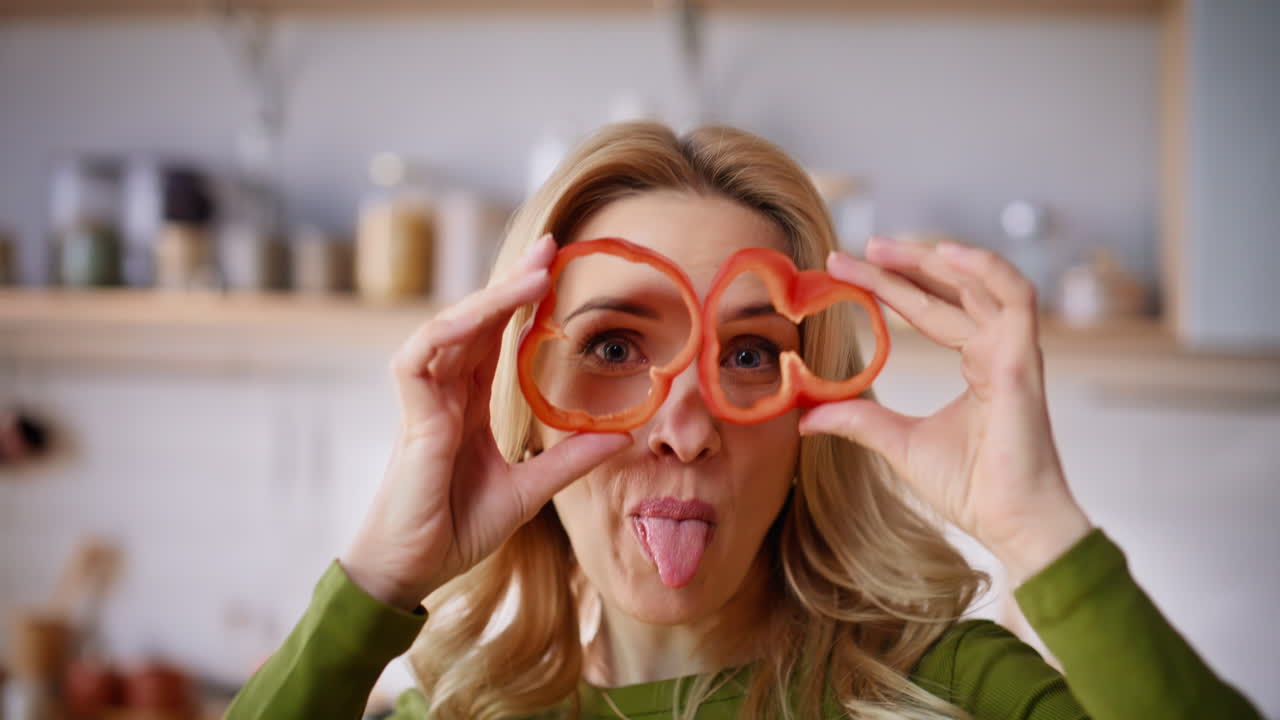 Ritratto di donna divertita che tiene fette di peperone fingendo che siano occhiali in una cucina luminosa.