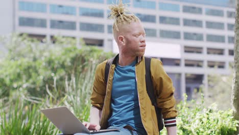 Thoughtful-albino-african-american-man-with-dreadlocks-sitting-in-park-using-laptop