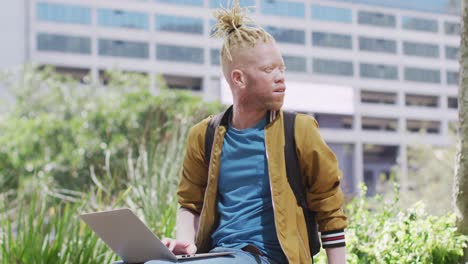 thoughtful albino african american man with dreadlocks sitting in park using laptop