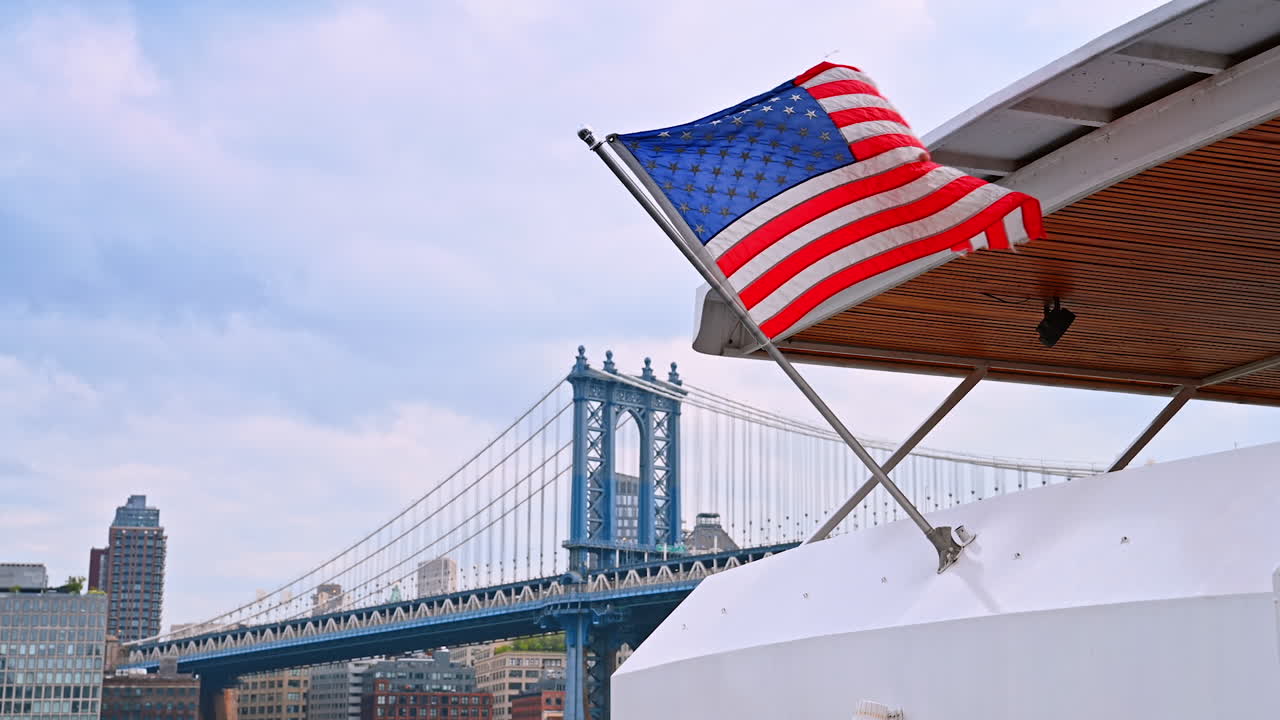 Ponte di Brooklyn con bandiera. Una barca con una bandiera americana naviga sul fiume, con il ponte di Brooklyn che incombe sotto un cielo nuvoloso