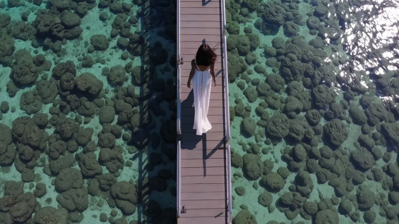 Woman in flowing white dress gracefully walks along wooden pier over vibrant coral reef, showcasing serene ocean beauty and tranquil exploration