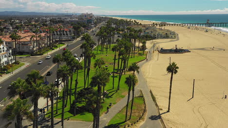 Drone-footage-flying-over-people-riding-bikes,-a-park,-palm-trees,-the-beach,-traffic,-with-a-view-of-the-Huntington-Beach-Pier