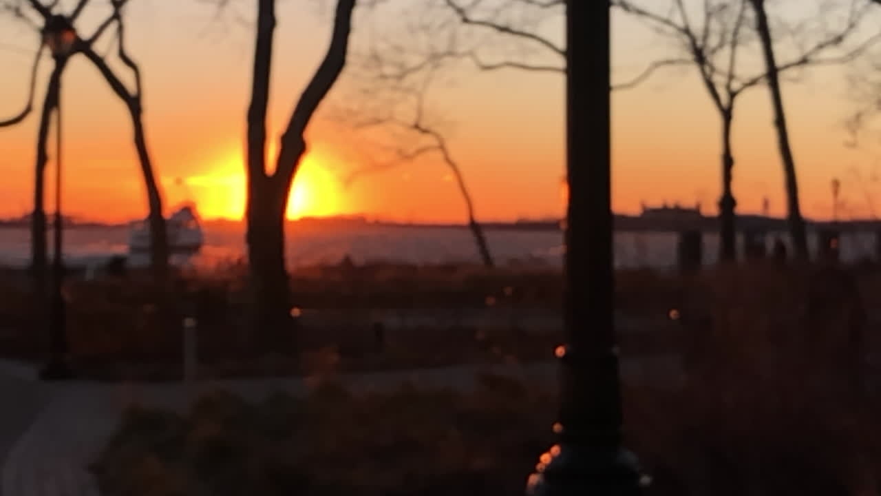 Sun going down in New York Harbor.Slow motion move through Battery park, sun reveals from behind trees and lamp post. Soft focus, lens flare.