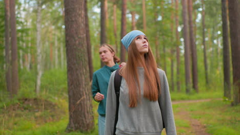 hikers walking along forest trail, woman in blue bandana and gray sweatshirt gazes around thoughtfully, while companion in green shirt adjusts collar