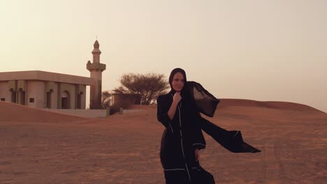 mujer musulmana de pie cerca de la mezquita en el desierto. viento fuerte oriente medio paz sin guerra