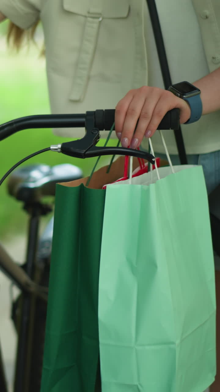 자전거 손잡이에 손을 고 산책하는 여성, 손잡이에서 매달린 녹색과 민트 쇼핑 가방, 그녀는 녹색 풀의 흐릿한 배경으로 검은색 크로스 보디 가방을 조정합니다.