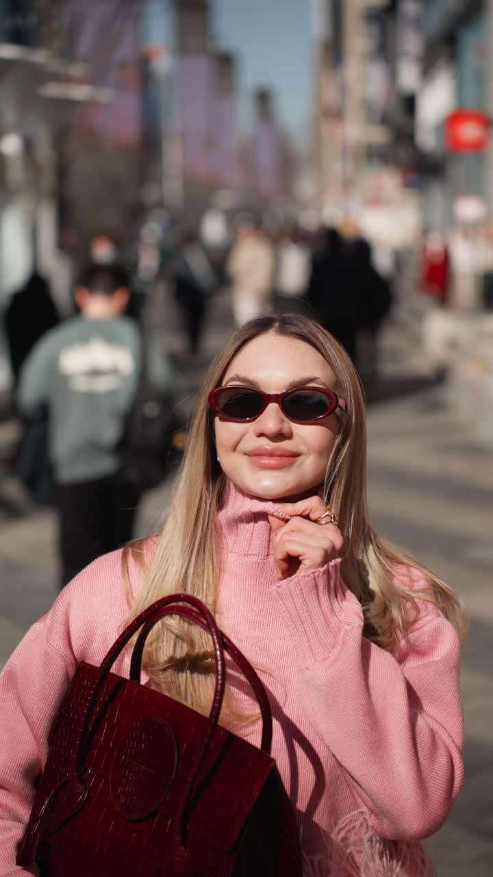Donna caucasica bionda e felice con borsa bordeaux che cammina su una trafficata strada cittadina in una giornata di sole - verticale.