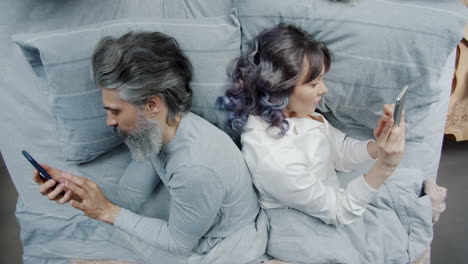 couple lying in bed, back to back, using phones
