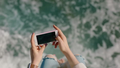 woman using smartphone taking photo of ocean legs dangling over water barefoot sharing summer vacation on social media watching waves