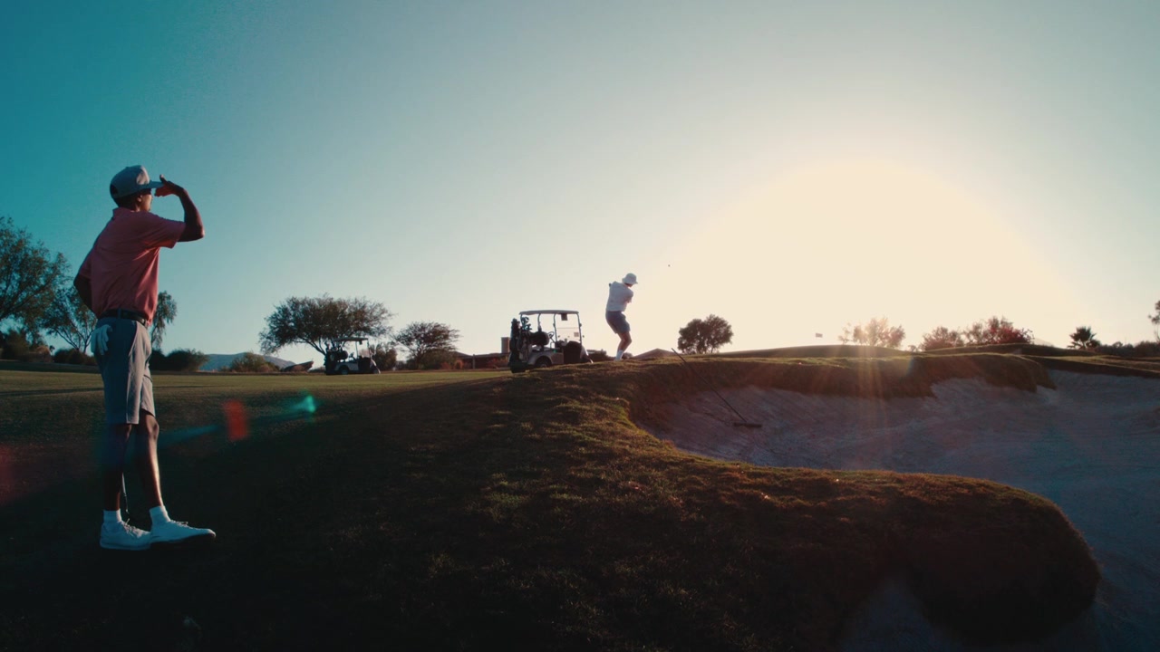 Un golfista osserva il suo compagno eseguire un pitch shot sopra un bunker mentre la luce del tramonto sagoma il campo desertico al rallentatore, ottimo per storie di lifestyle sul golf, pubblicità di resort e promozioni di viaggio