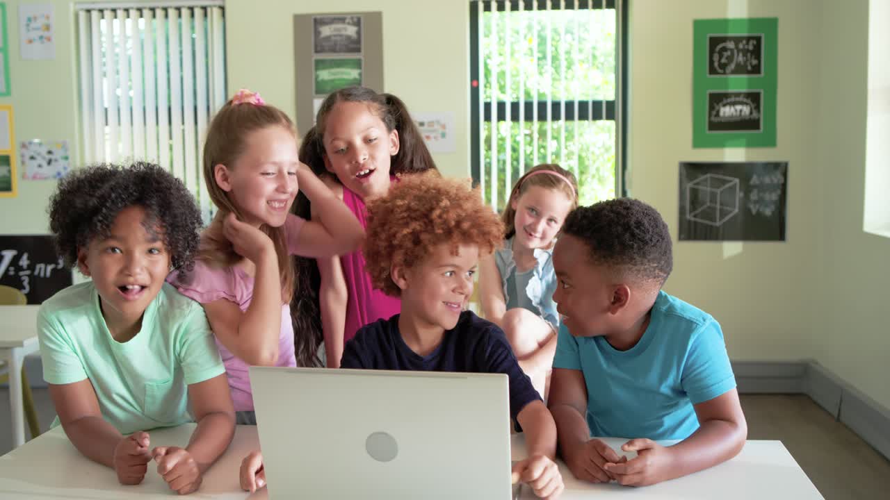 Bambini in età scolare di diverse etnie si radunano attorno a un computer portatile in classe, reagendo a una sorpresa sullo schermo.